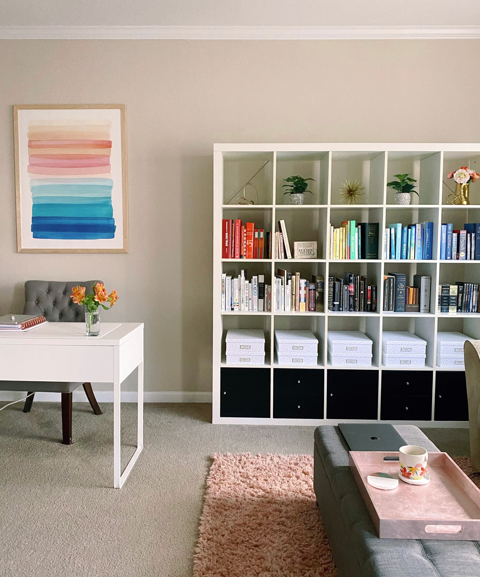living room with watercolor art on the walls, a simple desk with fresh flowers, and shelves full of color-coded books and simple decor