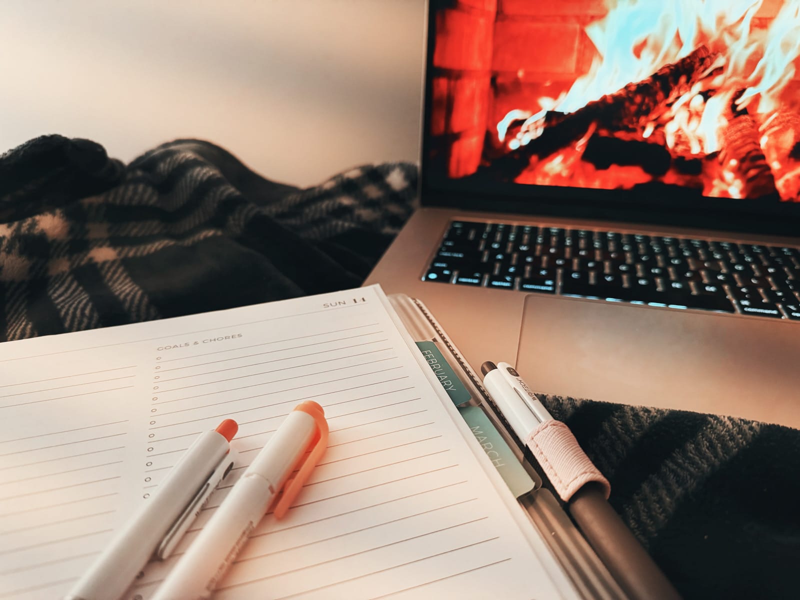 a paper planner (daily layout) with pens and highlighters nearby, as well as a faux fireplace video playing on a laptop screen in the background